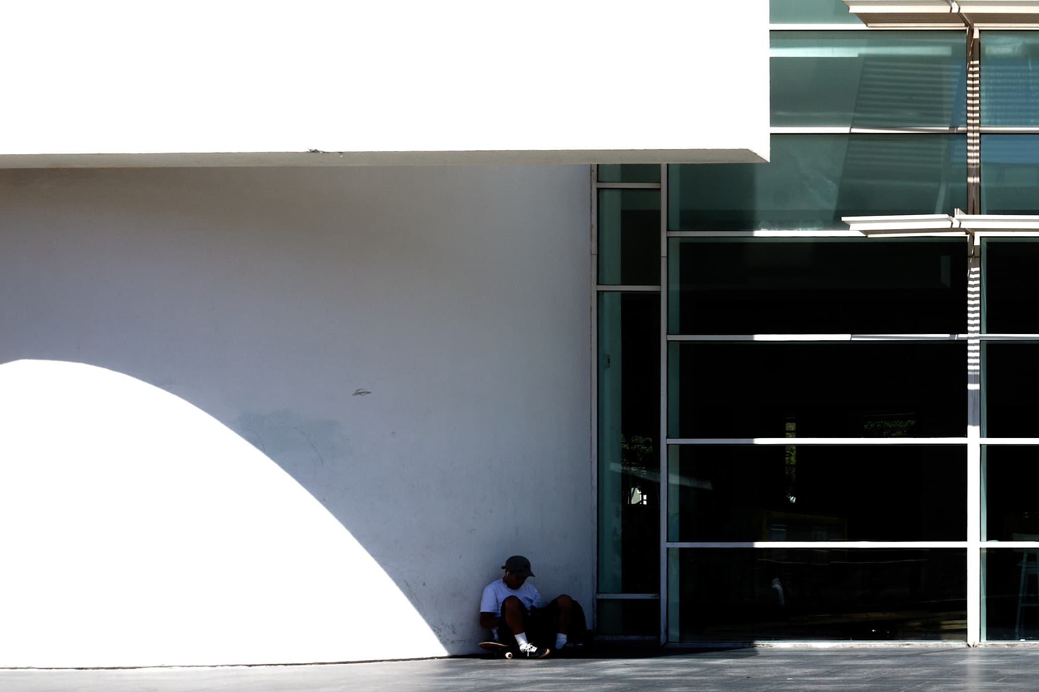 A skateboarder resting in the curved shadow of a modern building